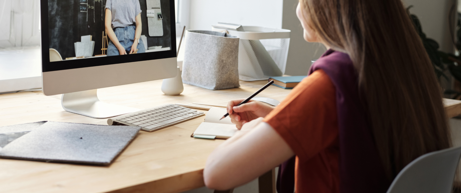 Kind sitzt mit Noitzblock am PC und schaut ein Video von einer Frau in der Schule