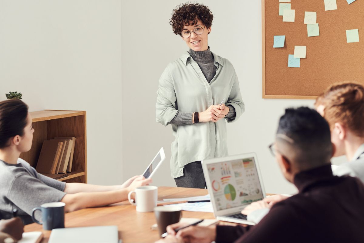 Frau steht vor Tafel mit weiteren Lehrkräften im Raum